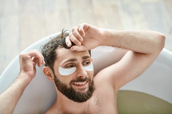 Homem bonito alegre com barba relaxante em sua banheira com manchas nos olhos, saúde mental — Fotografia de Stock