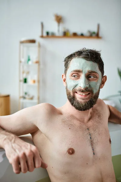 Gioioso bell'uomo con barba e maschera per il viso agghiacciante nella sua vasca da bagno, consapevolezza della salute mentale — Foto stock