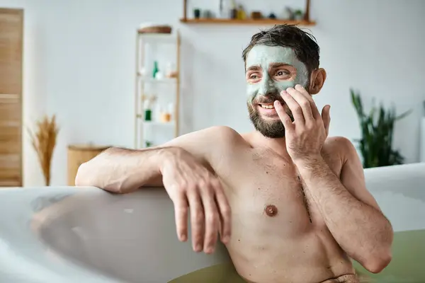 Gioioso bell'uomo con barba e maschera per il viso agghiacciante nella sua vasca da bagno, consapevolezza della salute mentale — Foto stock