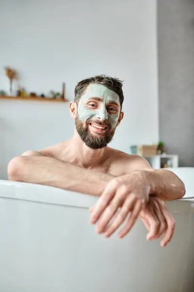 Gioioso uomo attraente con barba e maschera per il viso agghiacciante nella sua vasca da bagno, consapevolezza della salute mentale — Foto stock