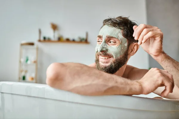 Fröhlicher attraktiver Mann mit Bart und Gesichtsmaske, der in der Badewanne chillt, Bewusstsein für psychische Gesundheit — Stockfoto