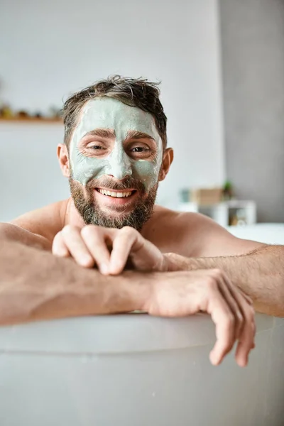 Радісний привабливий чоловік з бородою і маскою для обличчя, що охолоджується у ванні, обізнаність про психічне здоров'я — стокове фото