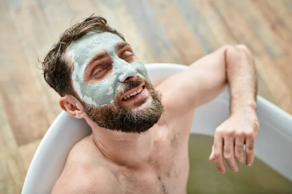 Симпатичный веселый мужчина с бородой и маской для лица, охлаждающийся в ванной, психическое здоровье — стоковое фото