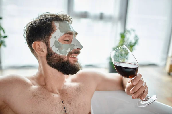 Весёлый привлекательный мужчина с бородой и маской для лица расслабляется в ванной с бокалом красного вина — стоковое фото