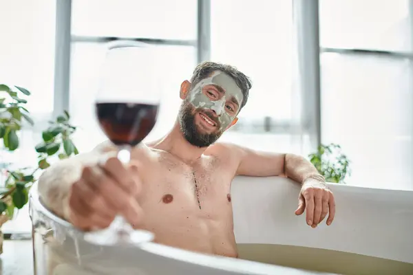 Fröhlicher attraktiver Mann mit Bart und Gesichtsmaske entspannt in der Badewanne bei einem Glas Rotwein — Stockfoto