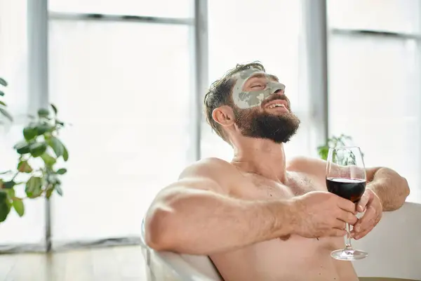Homem atraente alegre com barba e máscara facial relaxante na banheira com copo de vinho tinto — Fotografia de Stock