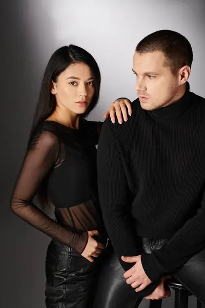 Verführerische asiatische Frau neben trendigem Mann in schwarzer Kleidung, der in die Kamera schaut, während er auf grau sitzt — Stockfoto
