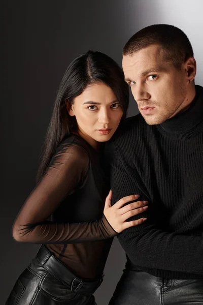 Appealing asian woman hugging handsome man in black clothes sitting on grey backdrop with light — Stock Photo