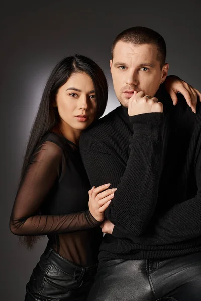 Fashionable expressive interracial couple in black clothes looking at camera on grey backdrop — Stock Photo