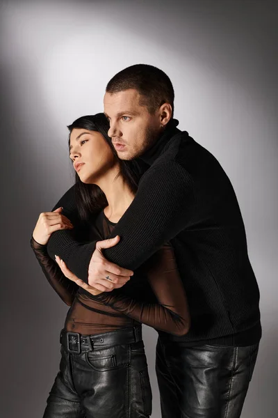 Leidenschaftlicher Mann in schwarzer Kleidung umarmt verführerische asiatische Frau vor grauem Hintergrund, Liebe und Mode — Stockfoto