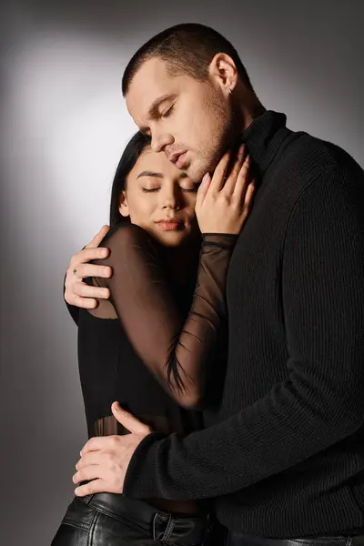Amante pareja interracial en negro ropa de moda abrazando con los ojos cerrados en gris telón de fondo - foto de stock