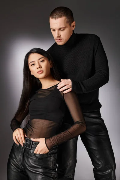 Séduisante femme asiatique en cuir noir pantalon regardant la caméra près de l'homme charismatique sur fond gris — Photo de stock
