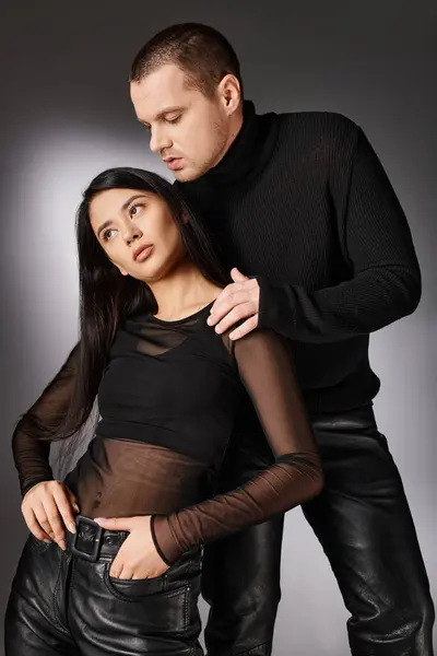 Passionate young man embracing brunette stylish asian woman in black clothes on grey backdrop — Stock Photo