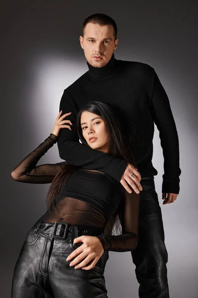 Handsome young man hugging brunette asian woman in black fashionable clothes on grey backdrop — Stock Photo