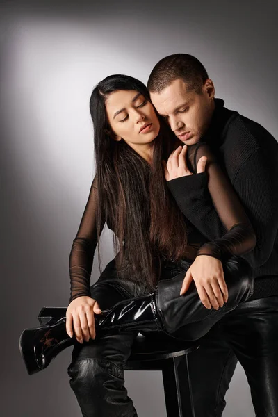 Jeune homme à la mode avec les yeux fermés embrassant femme asiatique à la mode assis sur fond gris — Photo de stock