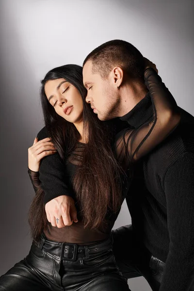 Romantic interracial couple in black casual attire embracing with closed eyes on grey backdrop — Stock Photo