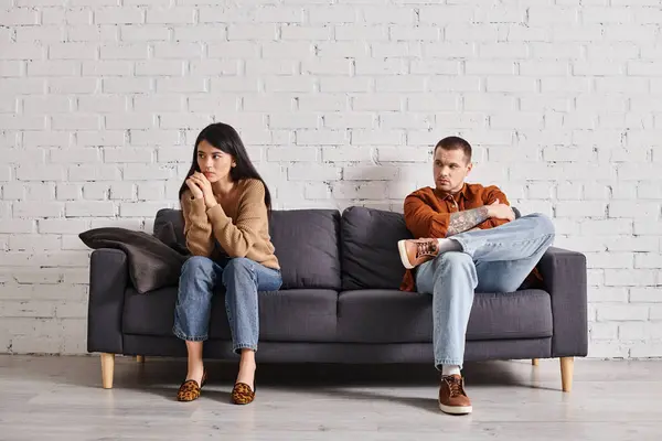 Young man looking at offended asian wife sitting on couch in living room, relationship trouble — Stock Photo