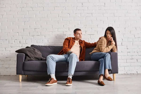 Giovane uomo calmante sconvolto moglie asiatica seduta sul divano in soggiorno, difficoltà di relazione — Foto stock