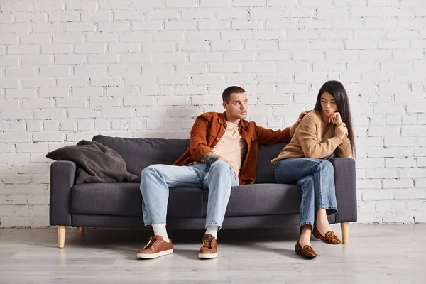 Young man calming upset asian wife sitting on couch in living room, relationship difficulties — Stock Photo