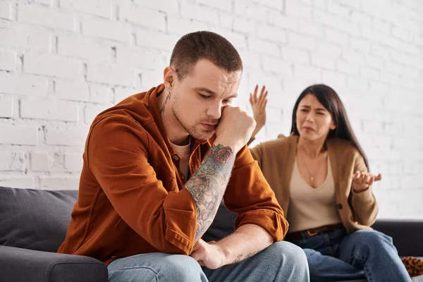 Irritated asian woman quarrelling at upset tattooed husband on couch at home, family problem — Stock Photo