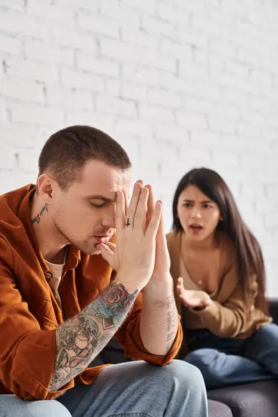 Jeune homme tatoué assis les yeux fermés près de l'homme asiatique en colère querelle à la maison, divorce — Photo de stock
