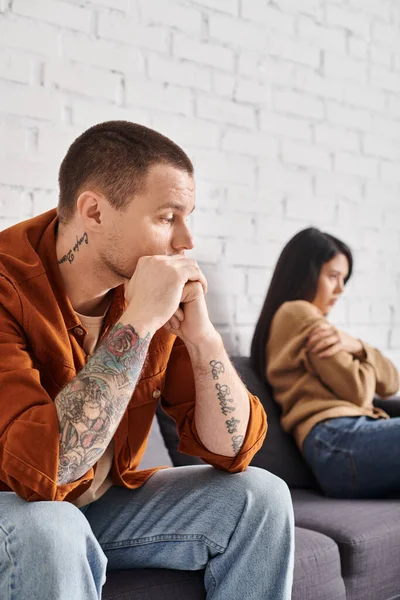 Frustrado tatuado homem olhando ofendido asiático esposa sentado no sofá em casa, divórcio conceito — Fotografia de Stock