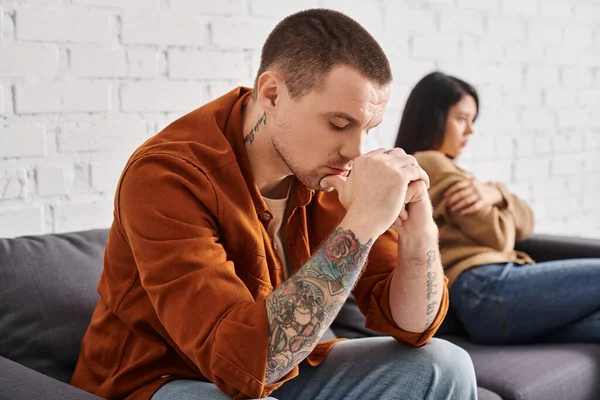 Depressed tattooed man with closed eyes near offended asian wife on couch at home, divorce concept — Stock Photo