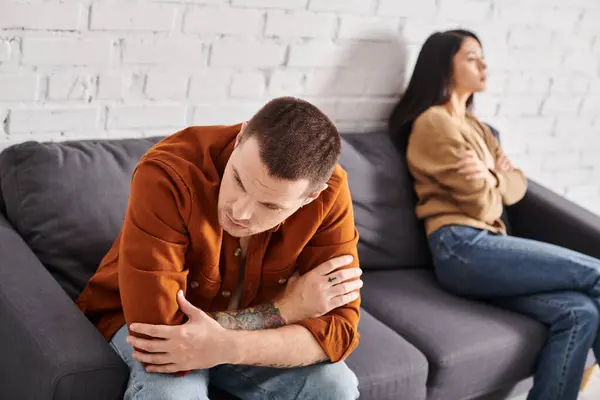 Похмурий татуйований чоловік сидить на дивані у вітальні біля ображеної азіатської дружини, сімейна проблема — стокове фото