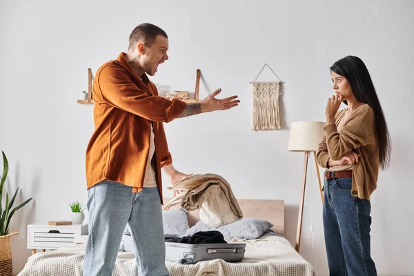 Junger und wütender Mann streitet mit asiatischer Frau in der Nähe von Koffer und Habseligkeiten im Schlafzimmer zu Hause — Stockfoto