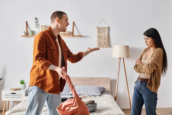 Young and angry man quarreling at asian wife near suitcase and clothes in bedroom at home — Stock Photo