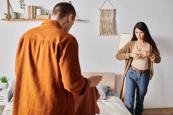 Offended asian woman taking off wedding ring near husband standing with clothes in bedroom, divorce — Stock Photo