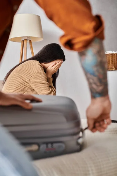 Tatuado hombre embalaje maleta cerca joven asiático esposa llorando en cama en casa, familia malentendido - foto de stock