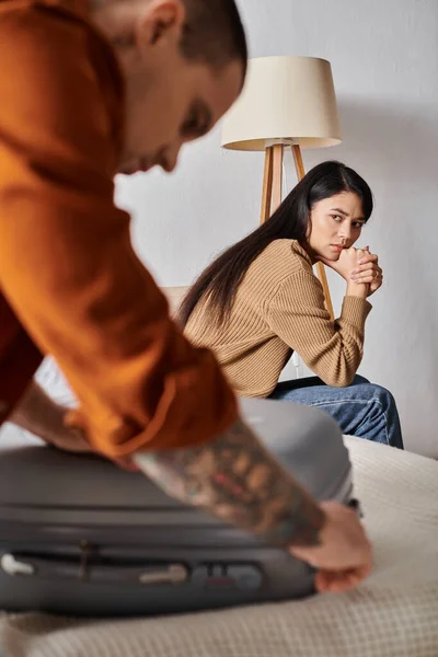 Gestresste Asiatin, die auf dem Bett sitzt und Ehemann beim Kofferpacken beobachtet, Scheidungskonzept — Stockfoto