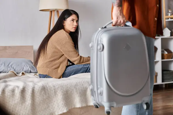 Verärgerte asiatische Frau sitzt auf dem Bett und sieht Ehemann beim Verlassen des Hauses mit Koffer, Scheidung — Stockfoto
