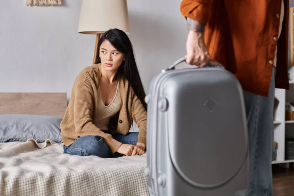Gloomy asian woman sitting on bed while husband leaving home with suitcase, divorce concept — Stock Photo
