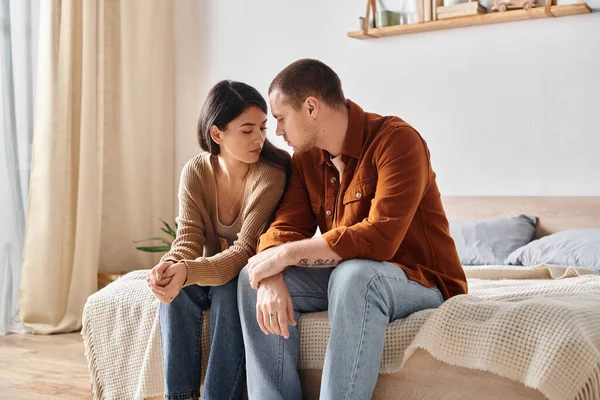 Joven y triste pareja interracial sentado y hablando en la cama en casa, dificultades de relación - foto de stock