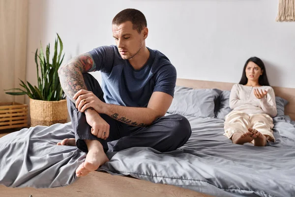 Upset tattooed man sitting on bed near frustrated asian wife, family relationship difficulties — Stock Photo