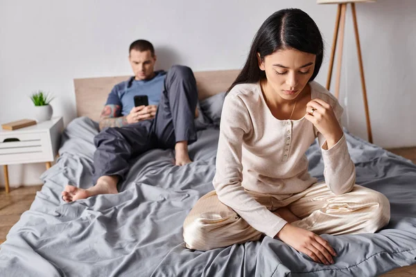 Offended asian woman sitting near husband using smartphone in bedroom at home, family conflict — Stock Photo