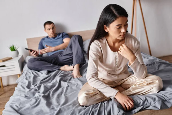 Frustrated asian woman sitting near husband using smartphone in bedroom at home, family problem — Stock Photo
