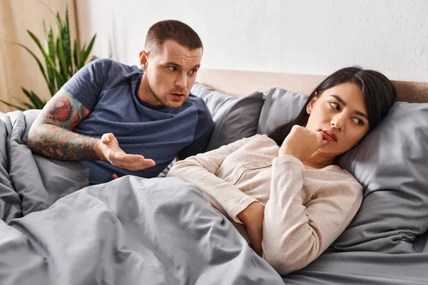 Irritated tattooed man quarrelling at upset asian wife on bed at home, family difficulties — Stock Photo