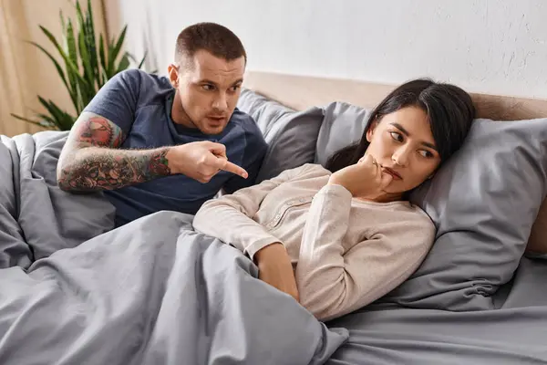 Angry tattooed man pointing and quarrelling at frustrated asian wife on bed at home, divorce — Stock Photo