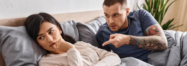En colère tatoué homme pointant et querelle à bouleversé asiatique femme sur lit à la maison, horizontale bannière — Photo de stock
