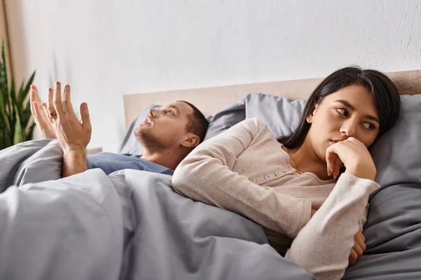 Young depressed asian woman lying down on bed near quarrelling husband, relationship conflict — Stock Photo