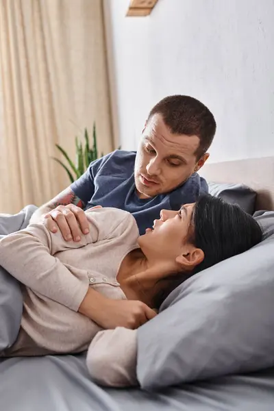 Joven marido tratando de calmar frustrado asiático esposa acostado en cama en casa, problemas familiares - foto de stock