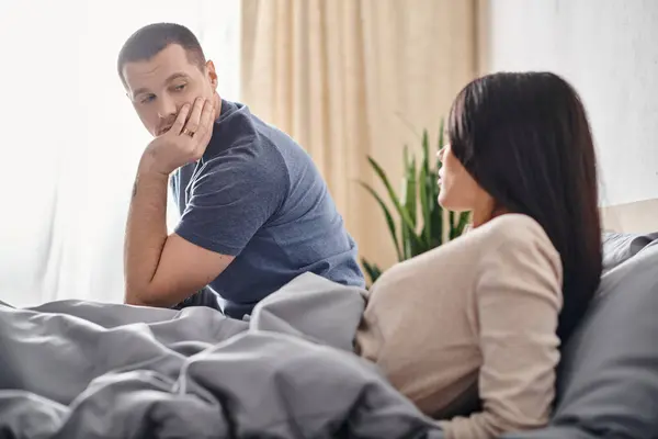 Young man talking to upset asian wife while sitting on bed at home, relationship difficulties — Stock Photo