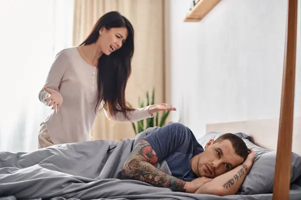 Irritated asian woman quarreling at offended tattooed husband lying on bed at home, divorce concept — Stock Photo