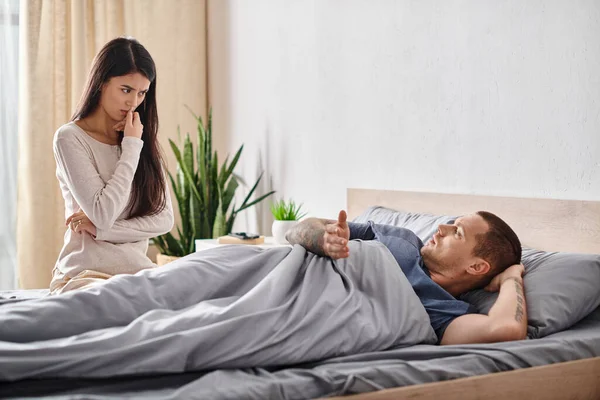 Ображена азіатська жінка, дивлячись на молодого татуйованого чоловіка, що лежить на ліжку вдома, концепція розлучення — стокове фото