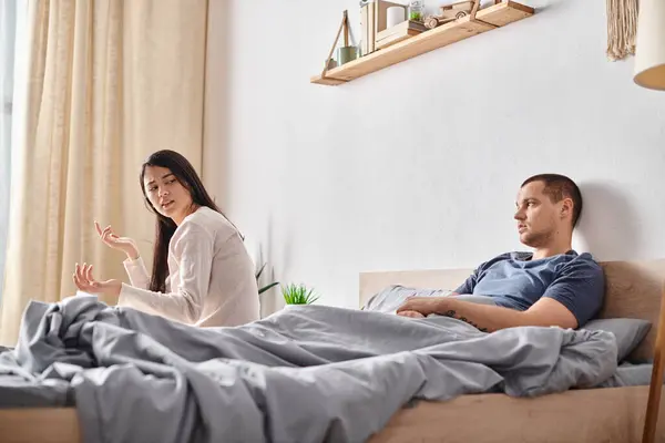 Offeso asiatico donna parlando con giovane tatuato marito sdraiato su letto a casa, divorzio concetto — Foto stock