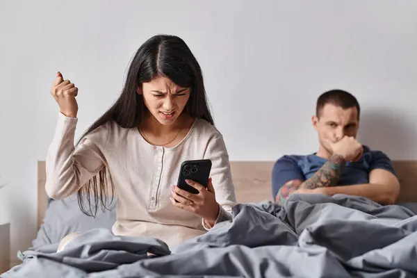 Angry asian woman looking at smartphone near husband lying on bed at home, family conflict — Stock Photo