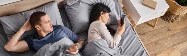 Uomo irritato litigando a giovane moglie asiatica guardando nel suo smartphone in camera da letto, banner — Foto stock